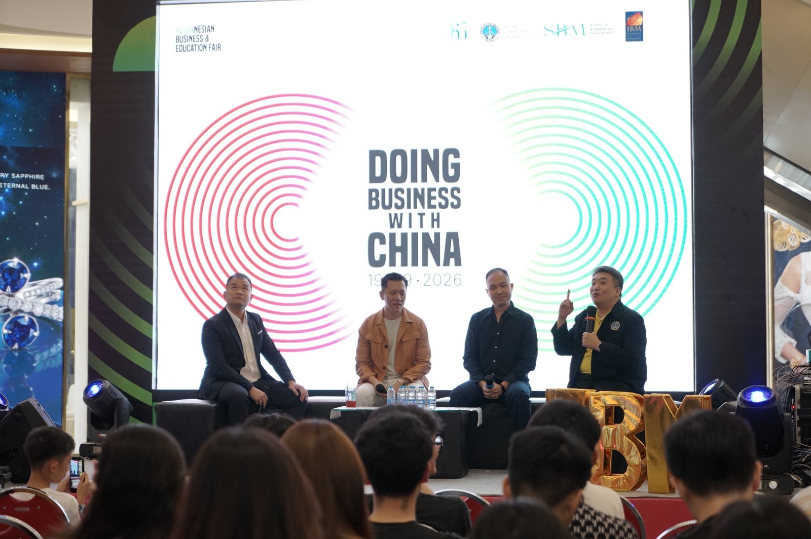 Four men seated on a stage in front of a large screen reading Doing Business With China, 19 09 2026, with gold IBM letters beside them and an audience watching from the front rows.