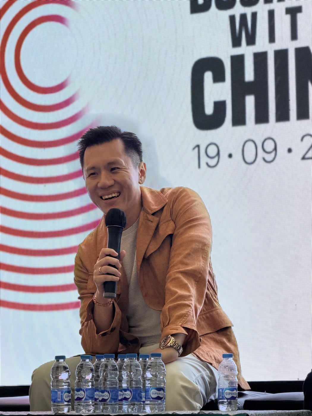 Stephanus Lukmanto smiling and holding a microphone on stage in front of the event backdrop.