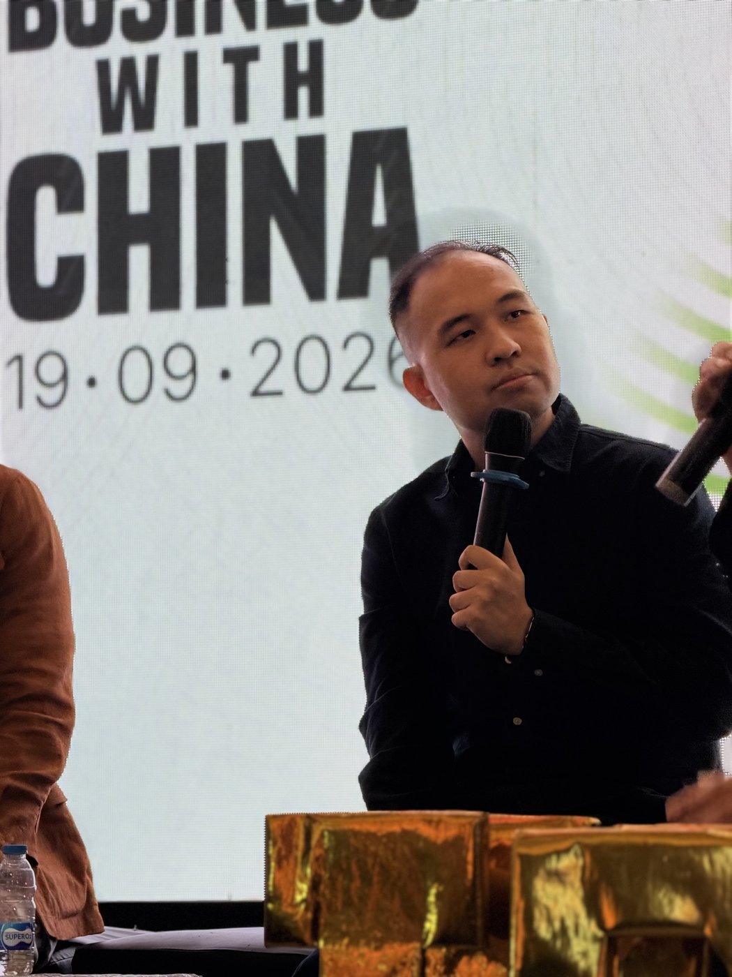 Billy Handoyo holding a microphone on stage in front of the Doing Business With China backdrop.