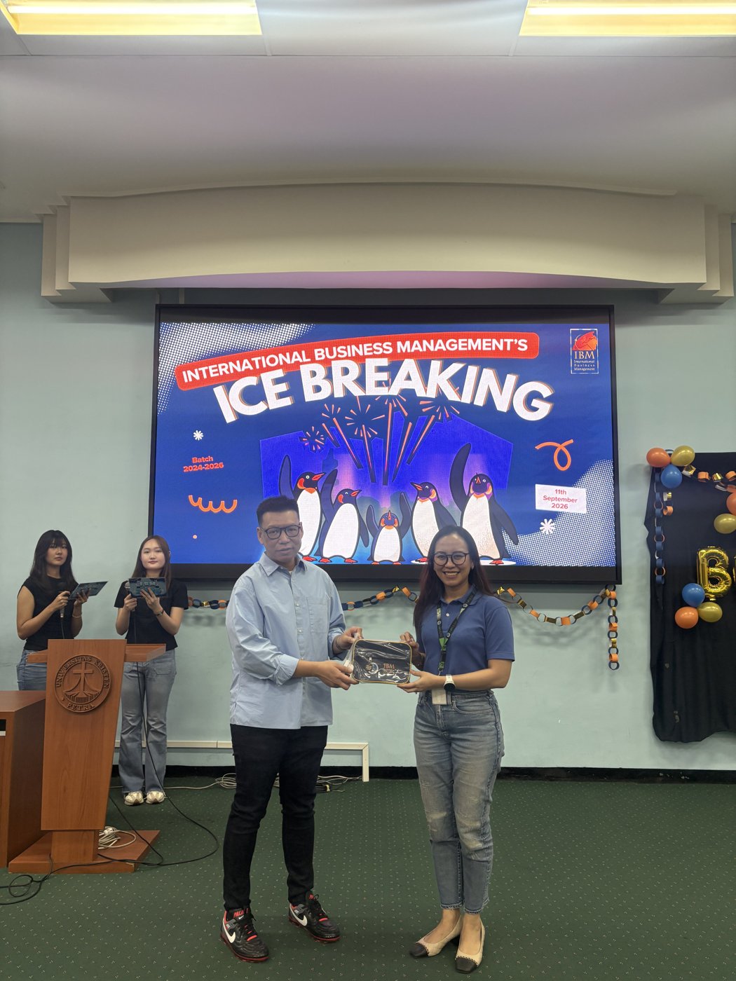 Mrs. Ruth Srininta Tarigan hands a token of appreciation to Hansen Thiono on stage, in front of a screen reading International Business Management's Ice Breaking, Batch 2024-2026, 11th September 2026.