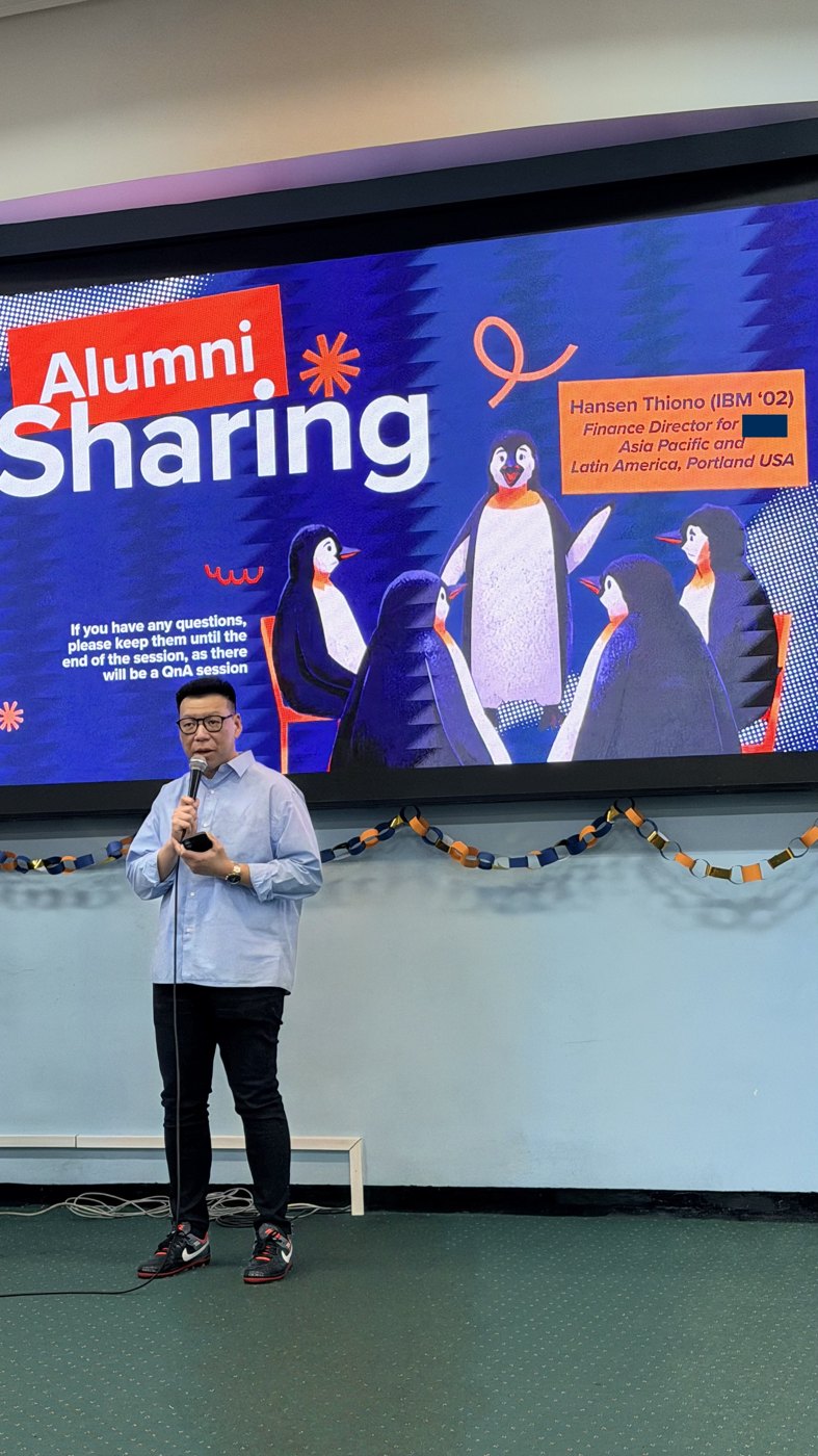 Hansen Thiono speaking into a microphone on stage, with the Alumni Sharing title slide on the screen behind him.
